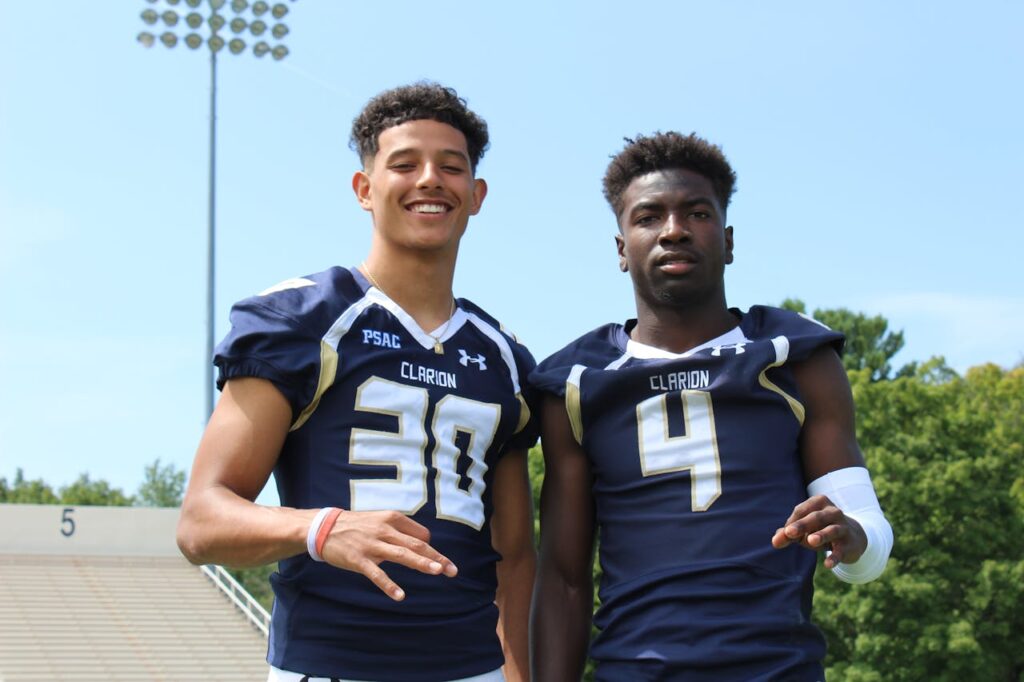 Two young football athletes posing in team uniforms outdoors on a sunny day.